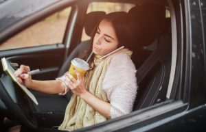 Woman sitting in her car, drinking a beverage, writing in a notebook and talking on the phone at the same time. 